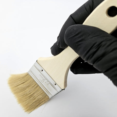 A person wearing a protective nitrile black glove holding a 2-inch GraphXOff™ Applicator Chip Brush with natural bristles, a metal ferrule, and wooden handle – Part #BB00013