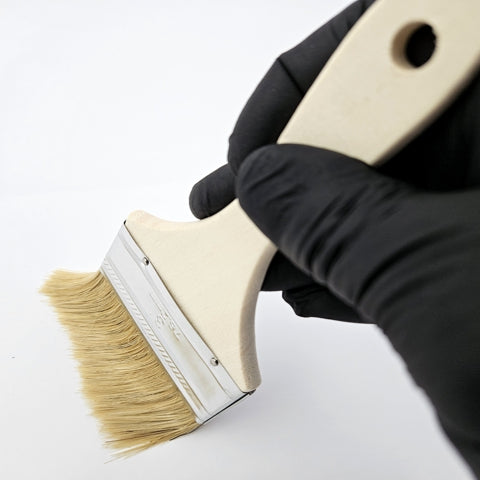 A person wearing a protective nitrile black glove holding a 3-inch GraphXOff™ Applicator Chip Brush with natural bristles, a metal ferrule, and wooden handle – Part #BB00015